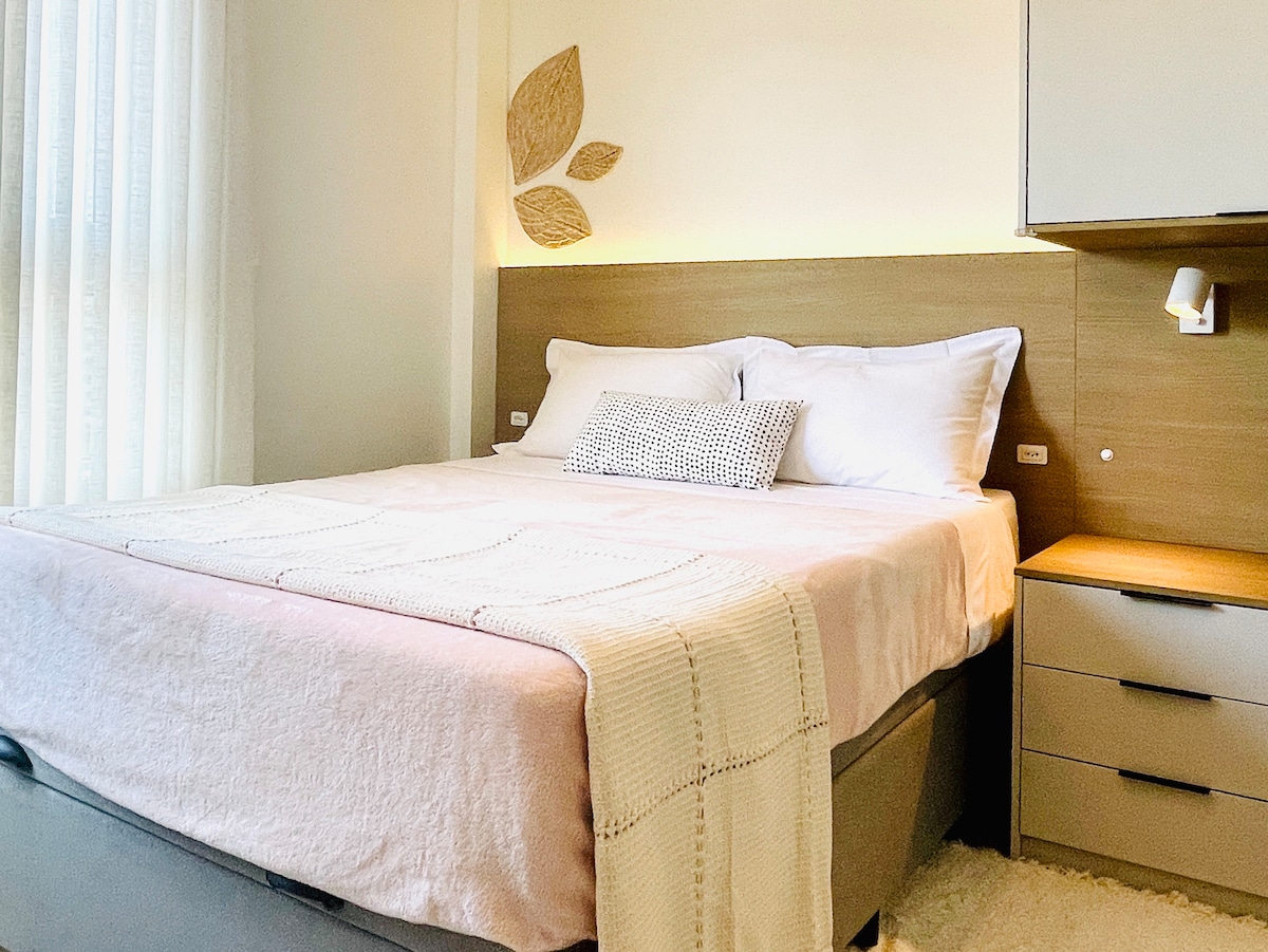 A cozy sleeping area features a spacious bed dressed with soft linens and a decorative pillow. Subtle lighting accentuates the headboard, while natural light filters through sheer curtains beside the bed. A bedside table holds essential items for convenience.