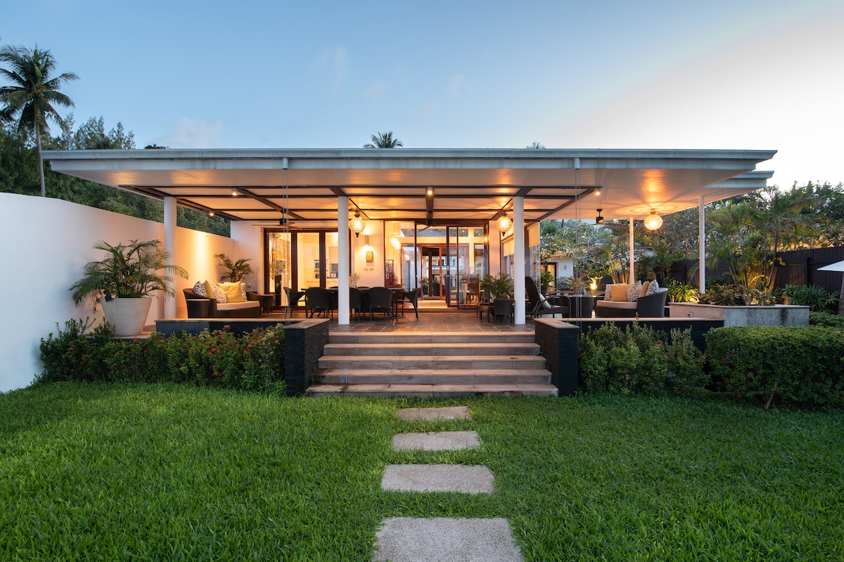 A modern villa exterior is set in a lush green garden, with stone steps leading to an inviting entryway. Large glass doors and windows enhance natural light, while pendant lighting creates a welcoming ambiance. Potted plants dot the outdoor seating area, contributing to a serene atmosphere.