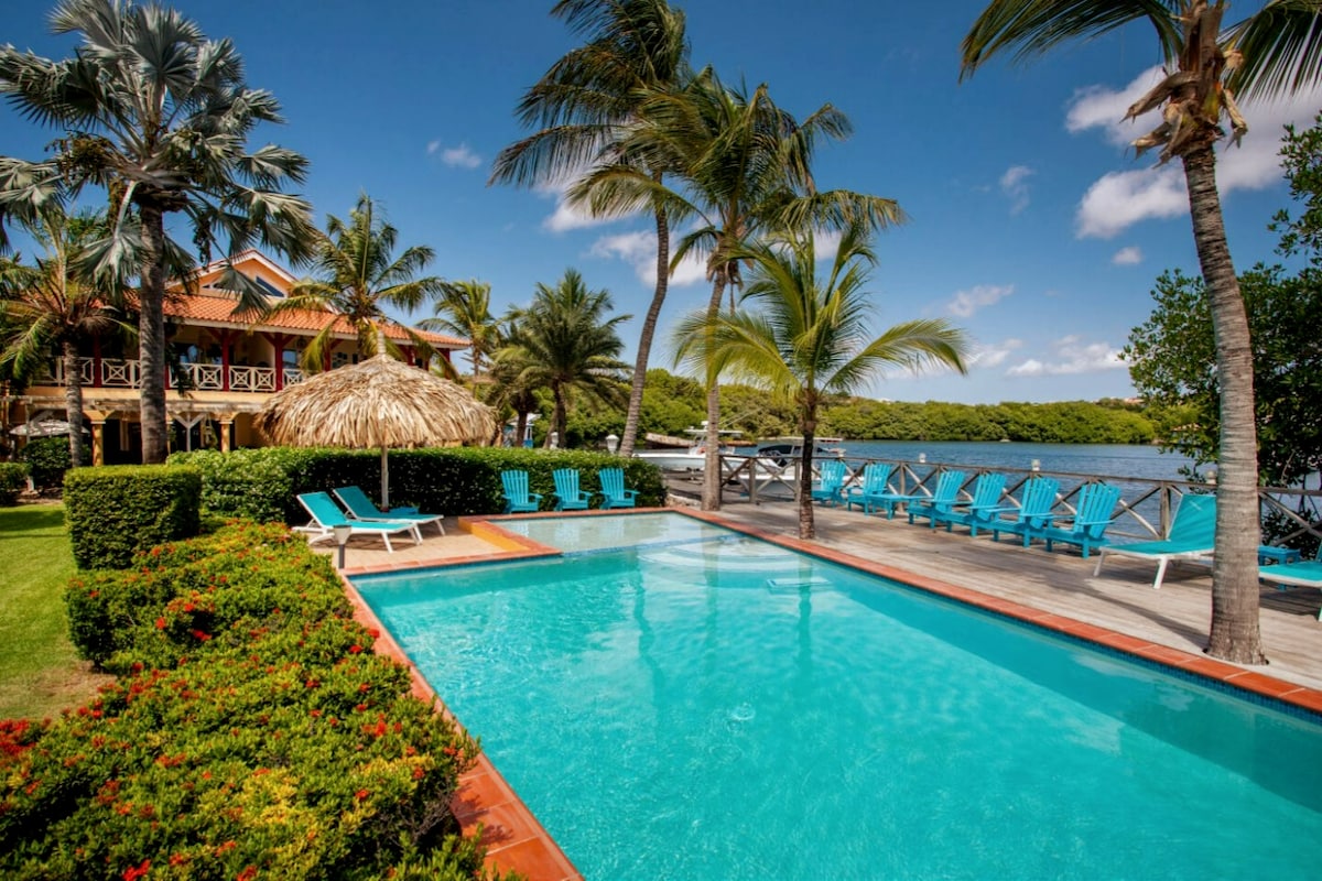 The image captures a serene outdoor scene featuring a large swimming pool surrounded by lush greenery and vibrant palm trees. Loungers in bright blue are arranged neatly by the poolside, while a private dock with seating is visible in the background, offering views of the tranquil water.