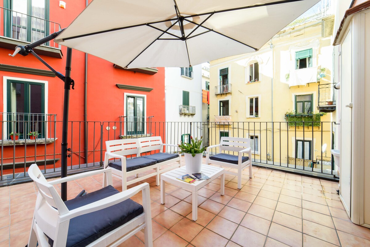 An outdoor terrace is featured with a white lounge set, including two chairs and a table. The area is shaded by a large umbrella, while colorful buildings surround the space, providing a vibrant urban backdrop.