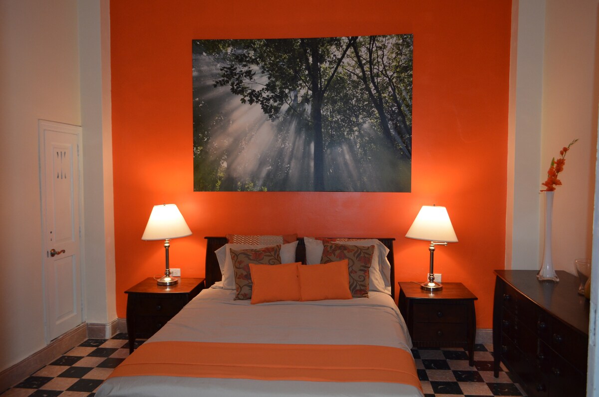 A spacious bed is centered in the room, adorned with a light-colored bedspread and vibrant orange accents. Flanking the bed are two wooden nightstands with lamps. A large artwork depicting trees and sunlight adorns the orange wall behind the bed, creating a warm ambiance.