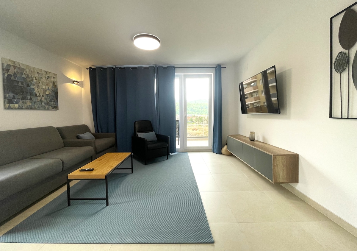 A spacious living area features a light grey sofa and an armchair, paired with a wooden coffee table. A flat-screen TV is mounted on the wall opposite. Large sliding doors provide access to a balcony, framed by long blue curtains, with views of the surrounding landscape.