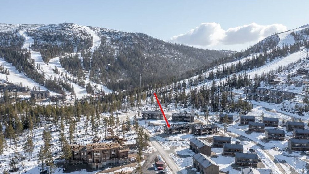 An aerial view captures the snow-covered landscape surrounding Klockarfjället. Ski slopes and residential buildings blend into the mountainous terrain, with a clear pathway leading to the featured apartment, highlighted by the red arrow. The serene winter scene emphasizes the proximity to outdoor activities.