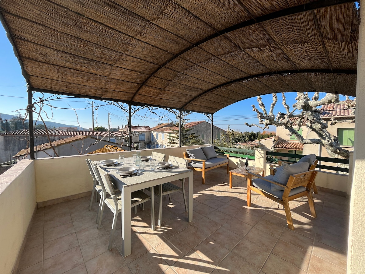 A spacious private terrace is covered by a thatched roof, featuring a dining table set with tableware and several comfortable seating options. The outdoor area is surrounded by greenery and offers views of the surrounding village rooftops under clear blue skies.