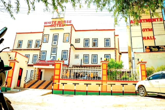The exterior of White Horse Hotel is displayed, featuring a multi-story structure with distinctive architectural details. Brightly painted with large windows, the building is framed by greenery and a low fence. An entrance leads to a staircase, indicating a welcoming approach.