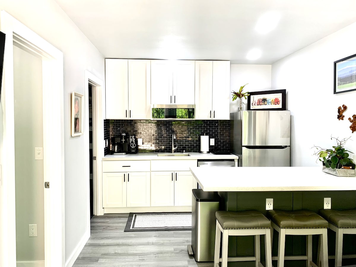 The sleek kitchen features white cabinetry complemented by dark tile backsplash. A stainless steel refrigerator stands beside a contemporary countertop with bar seating for four. Natural light streams in through a window above the sink, highlighting a well-organized and functional cooking space.