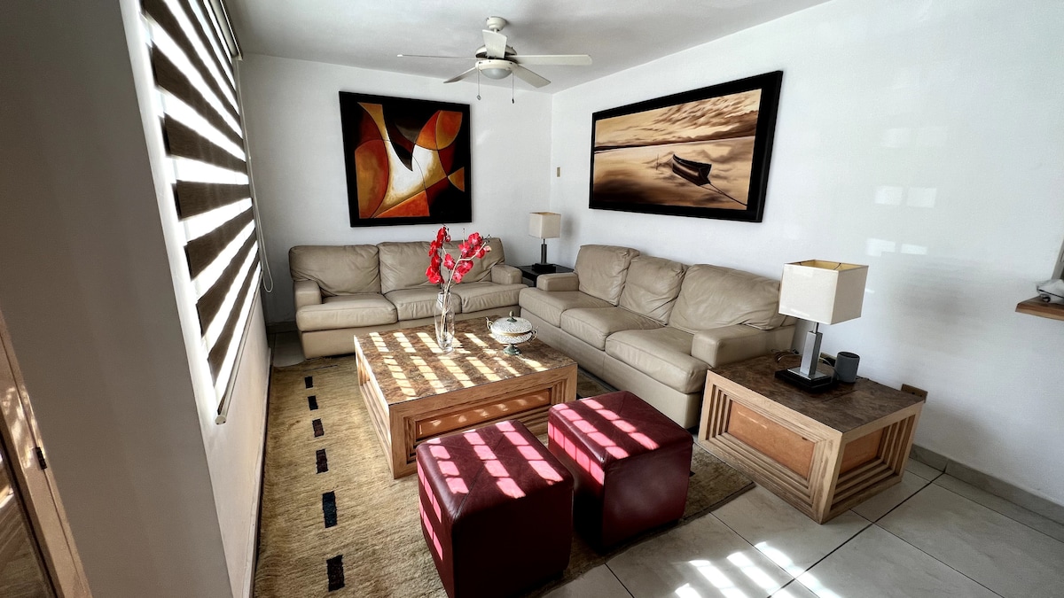 A well-furnished living area features two light-colored sofas arranged around a wooden coffee table. The space is decorated with vibrant wall art and illuminated by natural light filtering through the partially closed shades. Two red ottomans provide additional seating options.