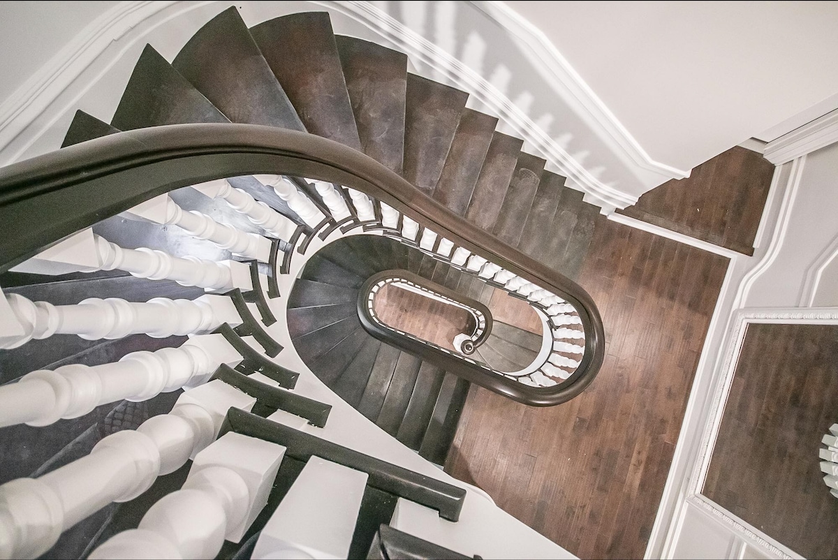 A spiral staircase is featured, showcasing a combination of dark wooden steps and white balustrades. The view from above highlights the elegant curves and contrasts of the staircase, which leads downwards to a polished wooden floor.