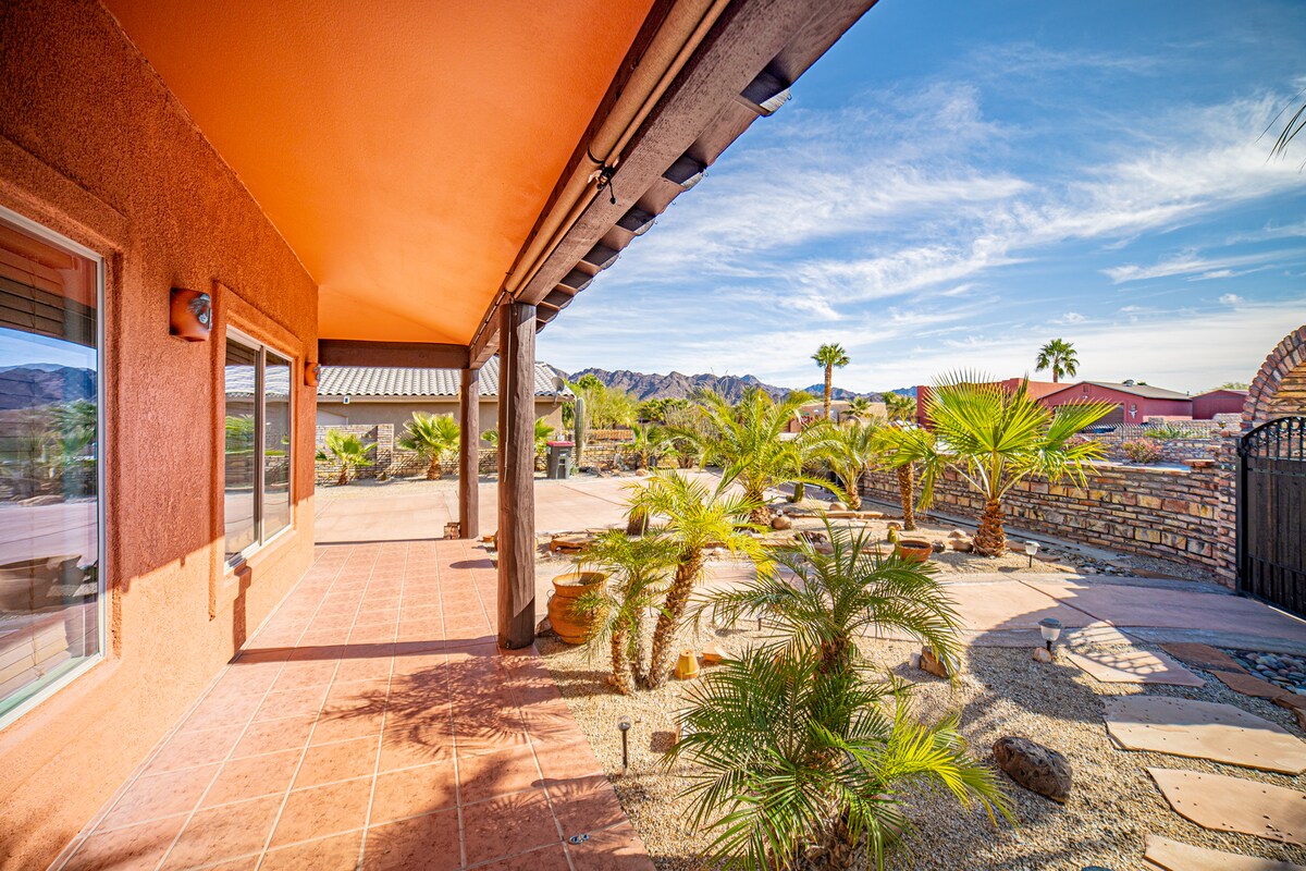An outdoor patio area is lined with vibrant palm trees and desert landscaping. Soft sunlight illuminates the terracotta tiles underfoot. The view includes distant hills and clear skies, while a stone wall adds a rustic touch to the space.