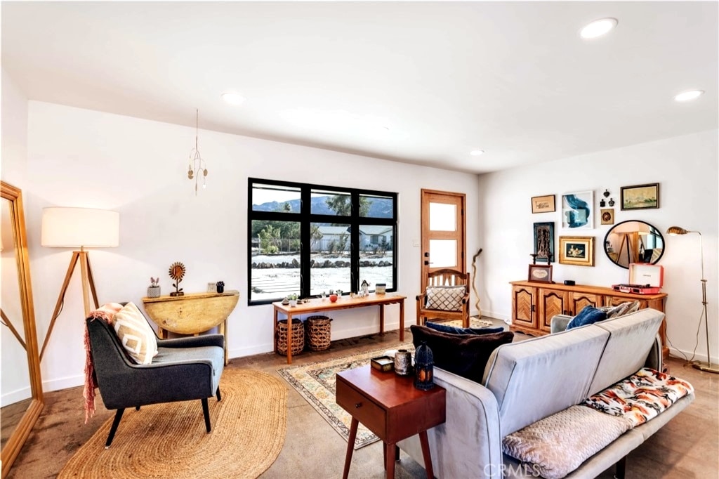 A bright and welcoming living room features large windows allowing ample natural light, with views of the outdoors. Comfortable seating is arranged around a central coffee table, and decorative accents are displayed on walls and surfaces, contributing to a calming atmosphere.