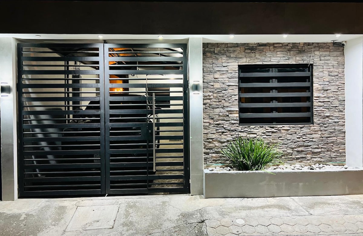 A modern exterior features a sleek, horizontal slatted gate that leads to a private garage, complemented by stone detailing on the wall. A small landscaped area with pebbles and greenery adds a touch of nature to the design.