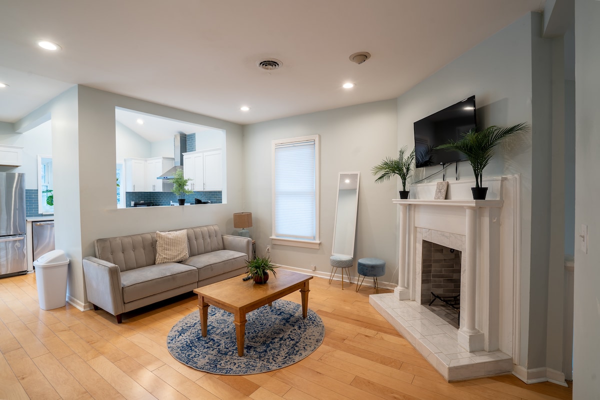An open living area features a light-colored sofa paired with a wooden coffee table. A decorative blue rug lies beneath the table. A fireplace is situated against a pale wall, while a large mirror and plants add subtle detail. Natural light brightens the room.