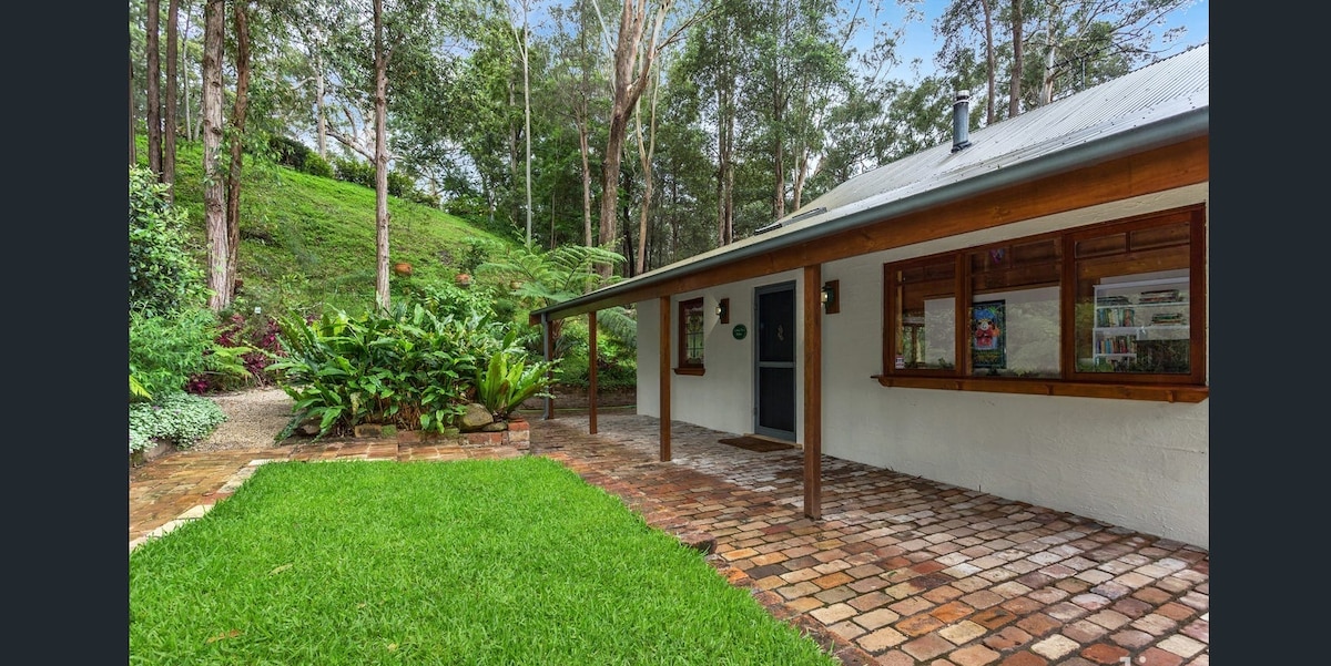 A charming cabin entrance is framed by a lush green lawn and native plants. The stone-paved terrace extends to the porch, providing access to large windows that invite natural light into the home, all set against a backdrop of towering eucalyptus trees.