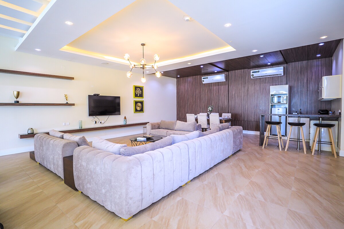 A spacious living area features a large sectional sofa arranged around a central low table. Wall-mounted shelves display decorative items, and a television is mounted on the wall. The kitchen area is equipped with modern appliances and bar stools provide additional seating.