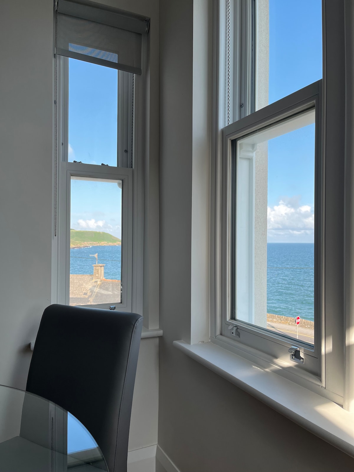Large windows frame a stunning view of the sea, allowing natural light to fill the room. A sleek dining chair is positioned beside a round glass table, creating a serene corner to enjoy the picturesque landscape outside.