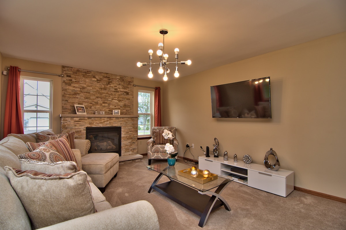A modern living room is displayed, featuring a cozy seating area with plush sofas and a stylish chandelier overhead. A stone accent wall is visible behind the fireplace, while a sleek coffee table and entertainment unit are positioned in front of a mounted flat-screen TV.