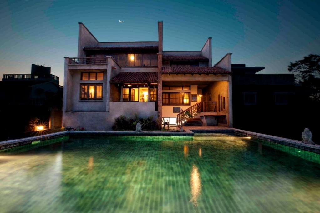 The villa's exterior is illuminated at dusk, showcasing a harmonious blend of modern architecture and traditional design. A large swimming pool reflects the surrounding soft lights, while the windows exhibit a warm glow, inviting guests to enjoy the space.