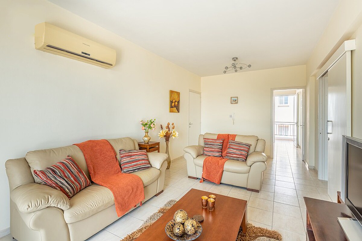The living area features two light-colored sofas adorned with orange throw blankets, arranged around a wooden coffee table. A decorative centerpiece with candles is placed on the table. The space is complemented by natural light entering through a nearby doorway.