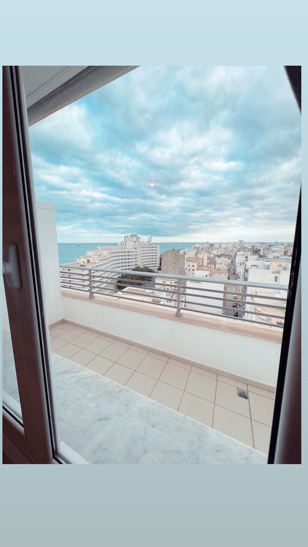 A view from a balcony showcases a sprawling urban landscape with buildings extending towards the horizon. The ocean is visible in the distance beneath a cloudy sky, with light reflections illuminating the scene. A railing surrounds the balcony area, complementing the serene atmosphere.