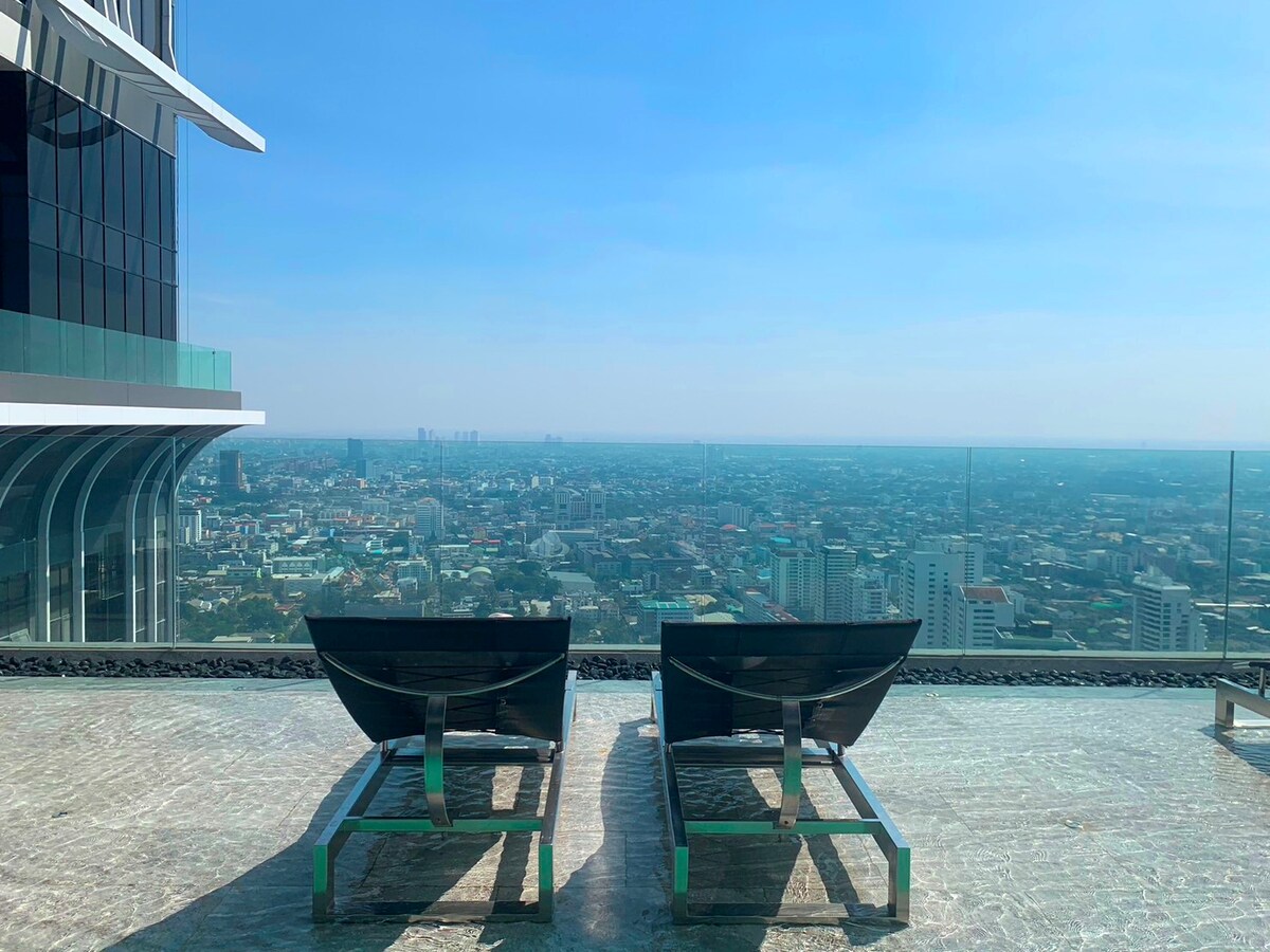 A rooftop terrace is featured with two lounge chairs positioned to overlook a panoramic city view. The space is surrounded by sleek glass panels, providing an unobstructed sight of the skyline under a clear blue sky.