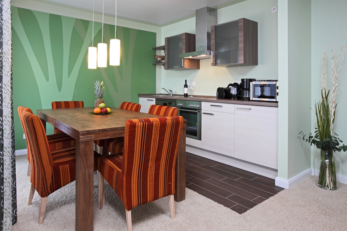 A modern kitchen area features a dining table surrounded by six upholstered chairs with bold stripes. The kitchen includes several appliances, such as an oven and microwave, against a soft green backdrop, while decorative elements like fruit are placed on the table.