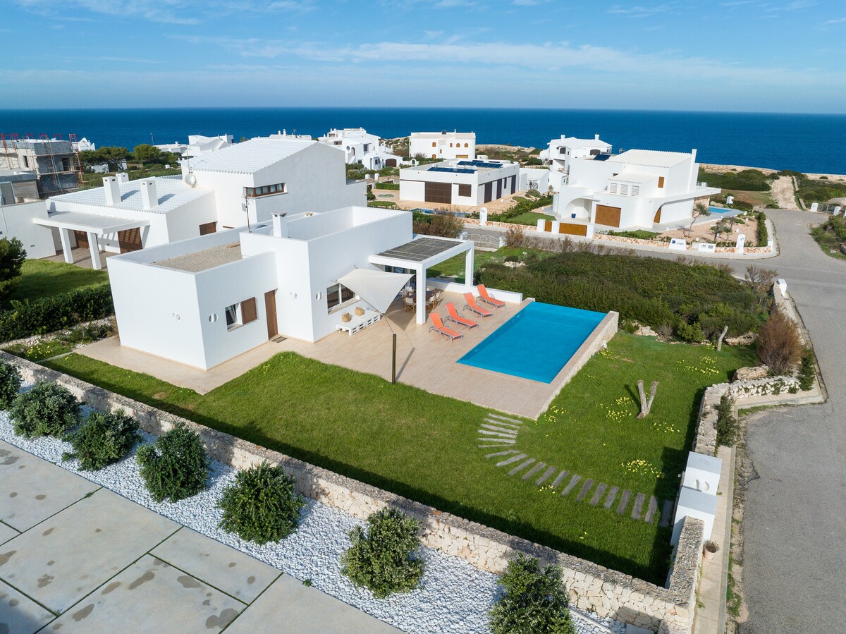 An aerial view of a modern villa is displayed, featuring a private swimming pool surrounded by a terrace with sun loungers. The well-manicured lawn leads to pathways and is bordered by decorative stone and shrubbery, with the stunning Mediterranean Sea visible in the background.