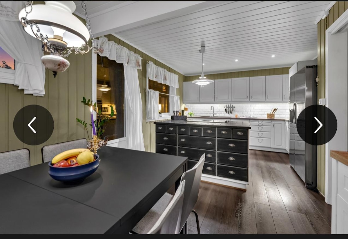 A well-equipped kitchen features a central island with black cabinetry and white countertops, offering ample workspace. Natural light enters through large windows, highlighting the dining area with a dark table surrounded by gray chairs. Decorative elements include a bowl of fresh fruit on the table.