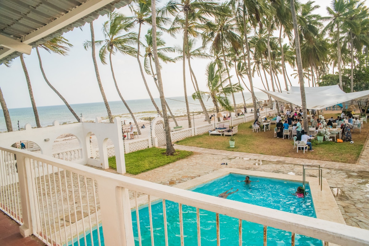 A refreshing blue pool is positioned within a sunlit area, surrounded by palm trees and a grassy lawn. In the background, a beachfront dining space is visible, filled with tables and chairs, while the horizon meets the serene ocean.