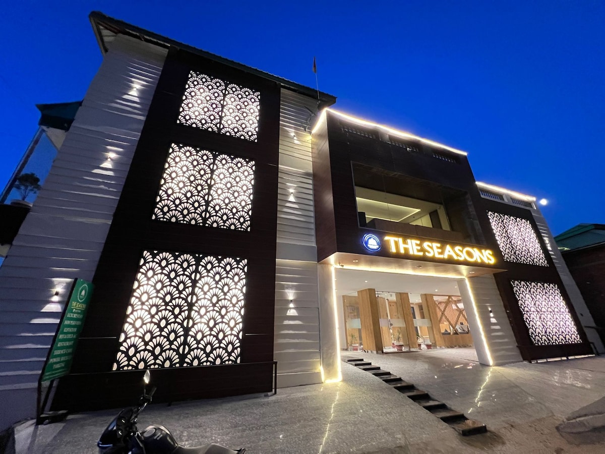 The exterior of the building is adorned with intricate latticework illuminated by soft lighting. The entrance features a welcoming sign displaying 'The Seasons'. A polished stone pathway leads to the inviting doorway, framed by modern architectural elements and a serene evening sky.