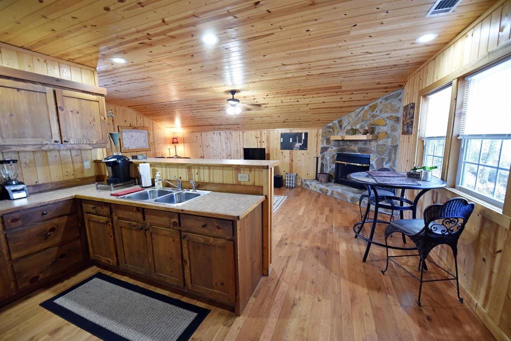 A warm kitchen area features wooden cabinets and a countertop with a coffee maker and sink. A circular dining table with two chairs is positioned beside a large window, while a stone fireplace adds a rustic touch to the inviting space.