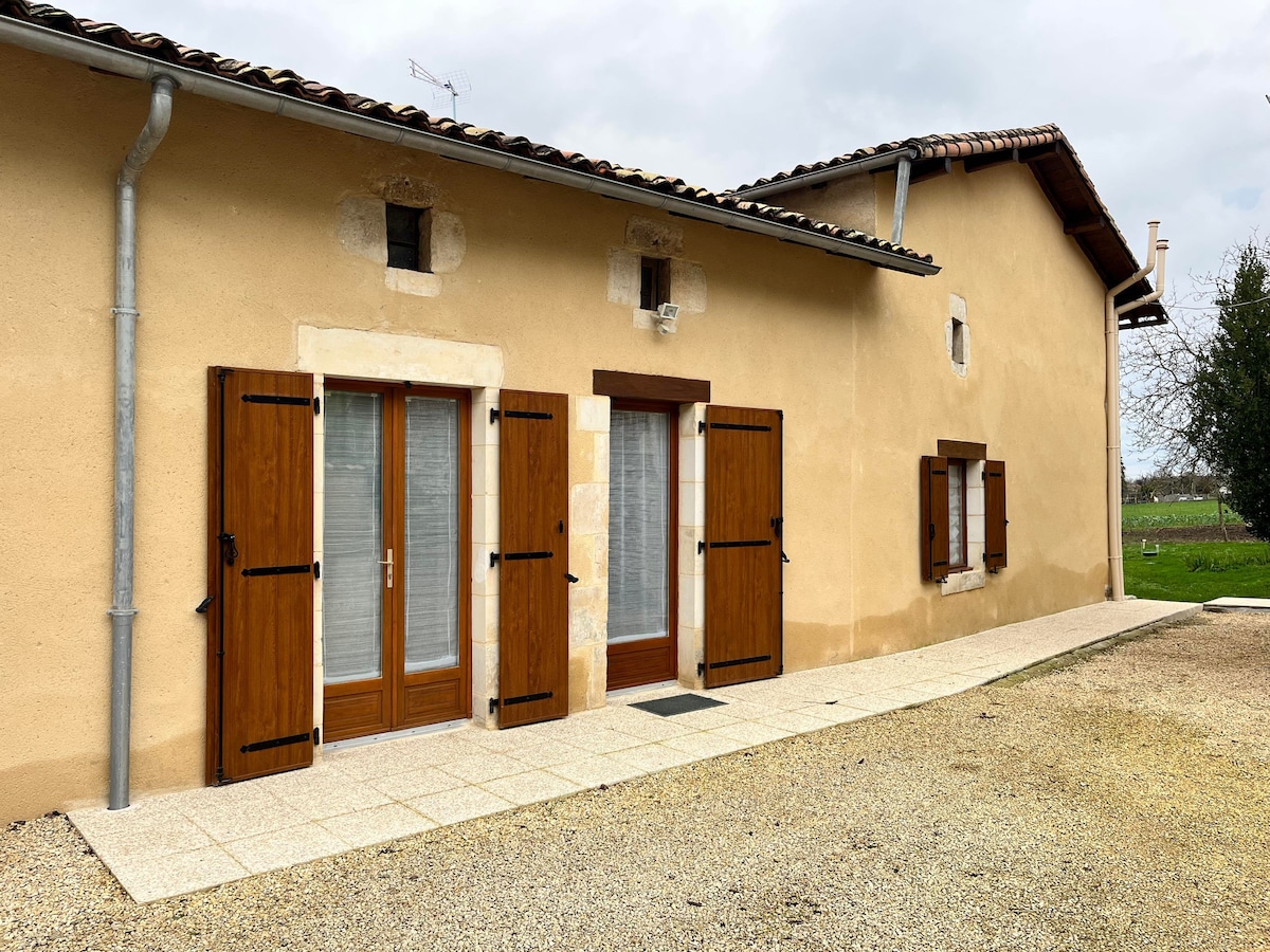 The exterior of a rustic home is presented, featuring natural stone walls and wooden shutters. A small stone pathway leads to the entrance, bordered by gravel. Large windows are framed by wooden accents, giving a sense of warmth and connection to the surrounding landscape.