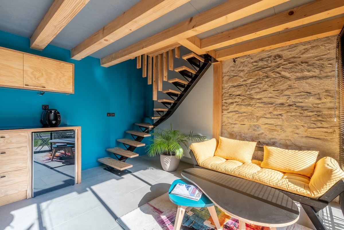 A bright living area features a yellow fabric sofa and a dark coffee table, inviting relaxation. Exposed wooden beams and a stone wall add texture, while stairs lead to an upper level. A plant adds a touch of greenery, enhancing the cozy ambiance.