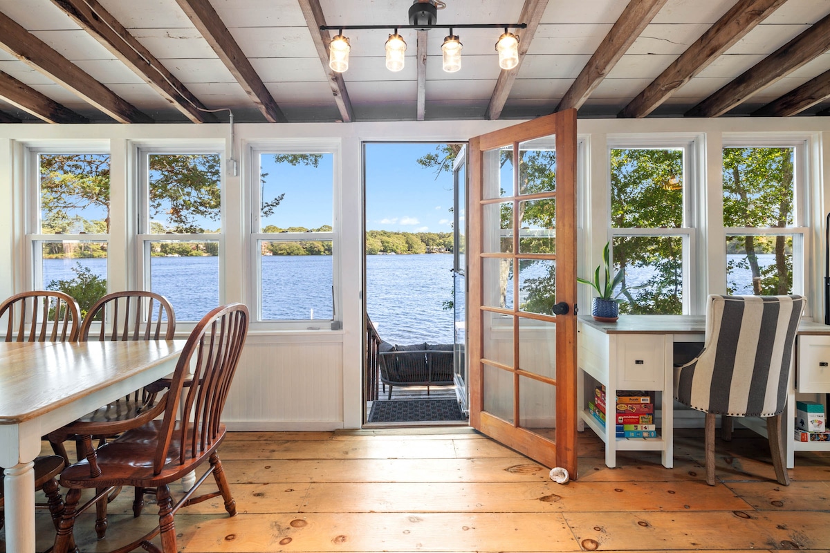 A view from the interior of the cottage showcases large windows framing the serene water scene outside. A wooden dining table with chairs is visible, along with a desk area that faces the water. Exposed beams line the ceiling, enhancing the rustic design.