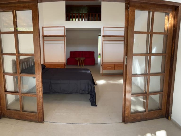 A spacious bedroom features sliding glass doors that open into the room. A dark bed is positioned against a neutral wall. A red sofa and a small wooden table are visible in the background, creating a comfortable seating area.