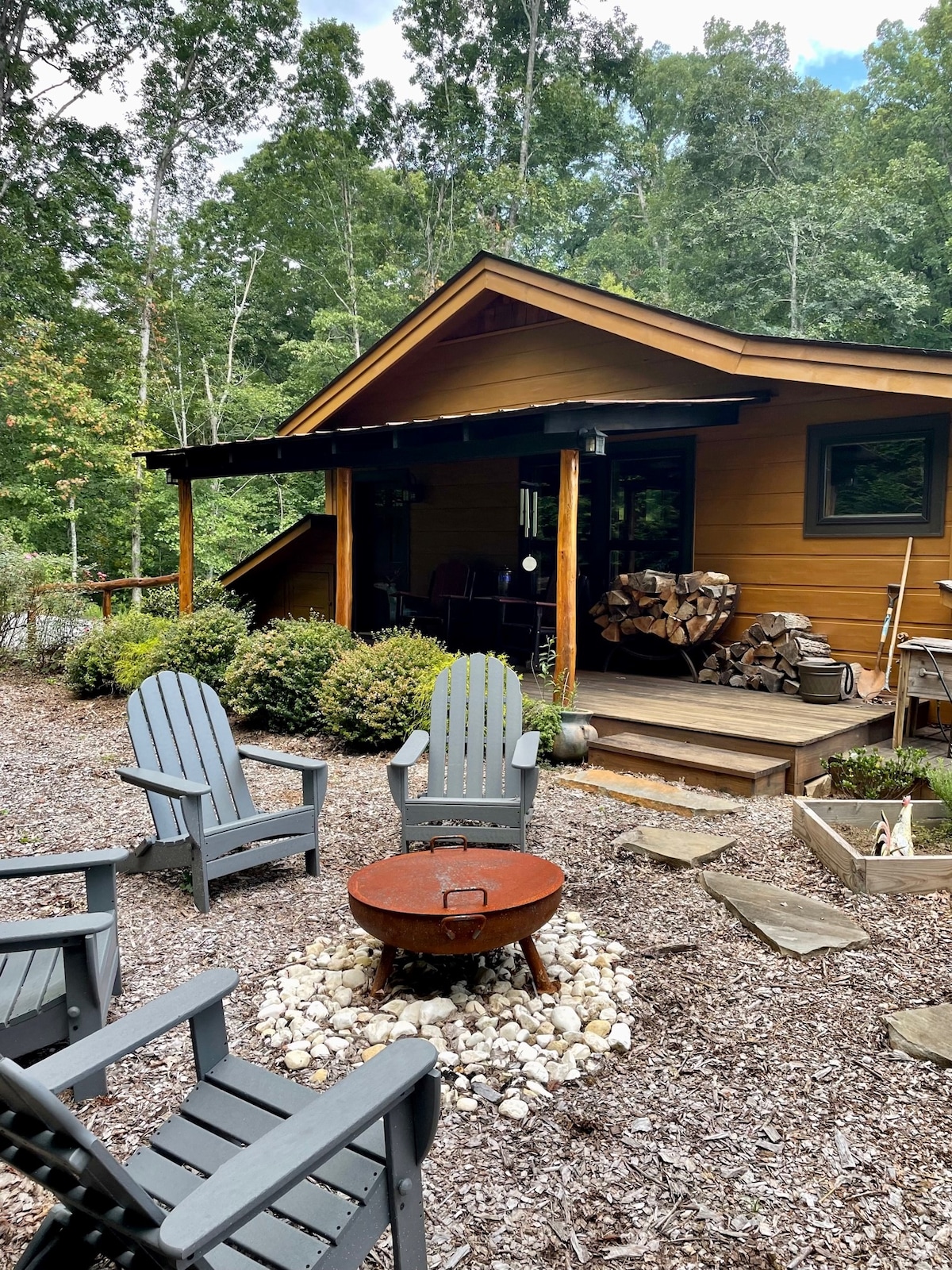 A cozy outdoor seating area is arranged with gray Adirondack chairs surrounding a stone fire pit. Wood is stacked nearby, and the guesthouse exterior features a rustic wooden facade, complemented by lush greenery and trees in the background.