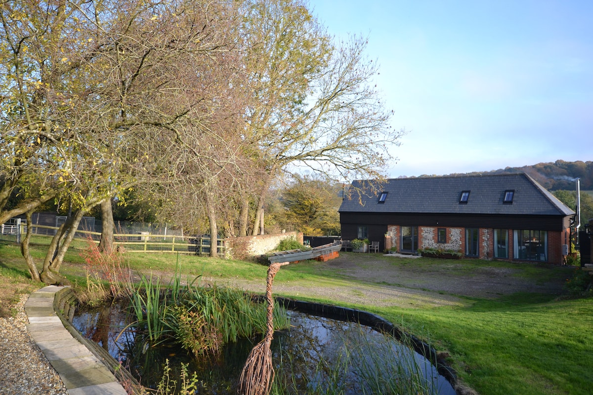 A serene view of the converted barn is presented, surrounded by lush greenery and a small pond. The landscape features distant hills and mature trees, creating a peaceful rural setting. A stone pathway leads towards the entrance, enhancing accessibility to the outdoor space.