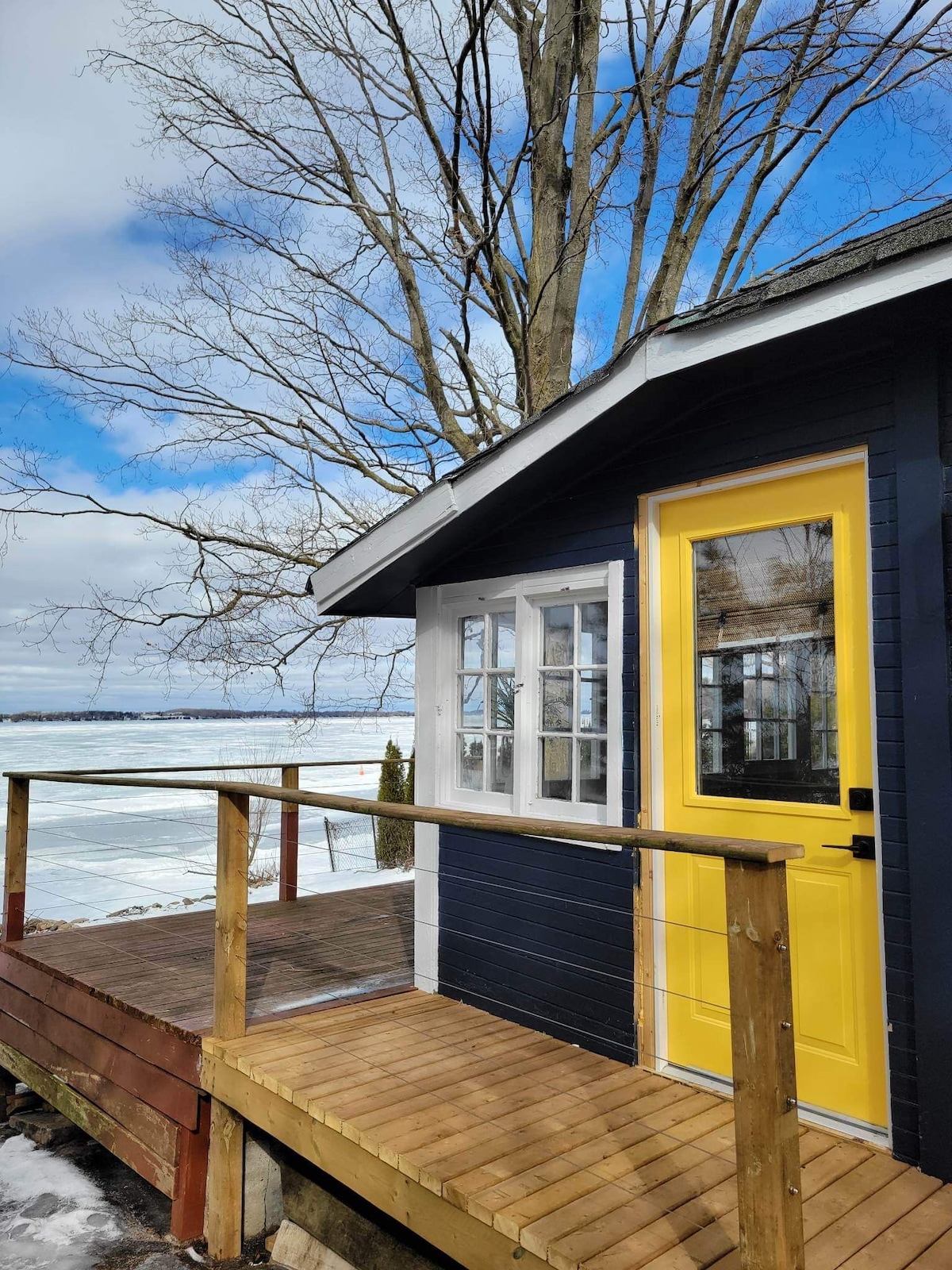 The cabin features a vibrant yellow door framed by large windows, allowing for natural light. A wooden deck extends from the entryway, offering a view of the icy lake and surrounding trees under a partly cloudy sky.