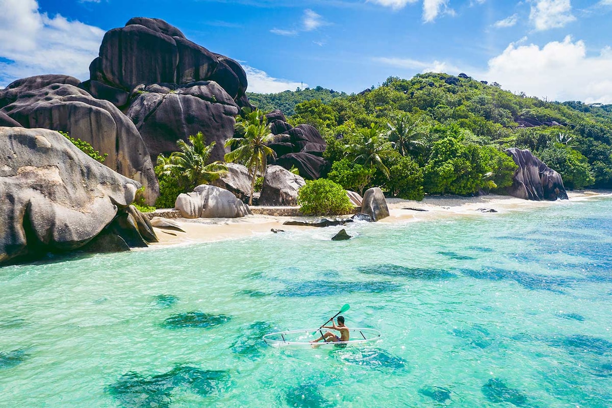 A kayaker glides across clear turquoise waters near a lush, green shoreline. Large granite boulders and palm trees frame the picturesque beach, highlighting the natural beauty of the area. The vibrant sky features scattered clouds, illuminating the scenic landscape.