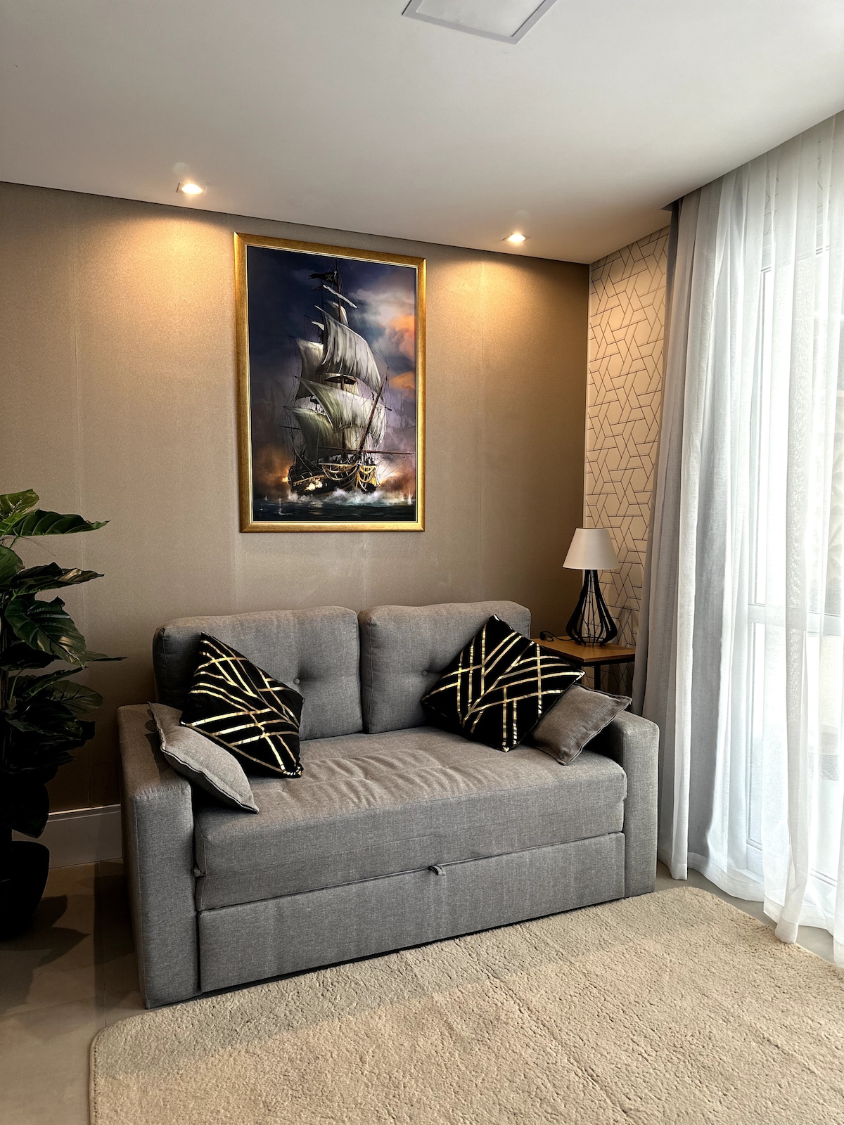A cozy seating area is presented, featuring a gray sofa adorned with decorative cushions. A lampshade stands on a side table, complementing the framed artwork of a ship, which is displayed on the wall. Natural light filters through sheer curtains.