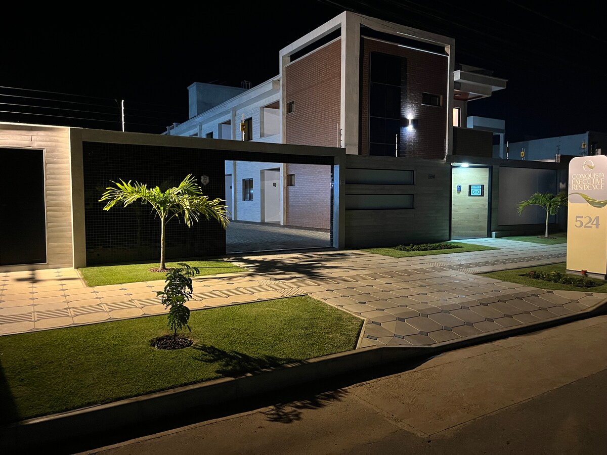 The exterior of the property is displayed at night, featuring a well-lit entrance and modern architecture. A manicured lawn with a small tree is visible, alongside a patterned walkway leading to the main entrance.