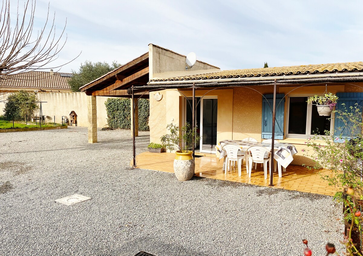 A tranquil patio area features a tiled floor, surrounded by a spacious gravel yard. A round table with several white chairs is positioned under a wooden overhang, with blue shutters framing the nearby glass doors that lead into the house.