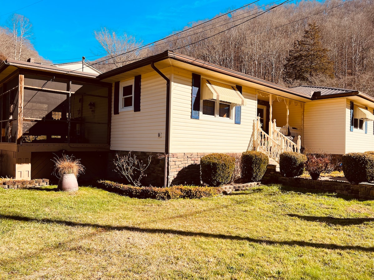 The exterior of the home is showcased, featuring a single-story structure with a welcoming entryway and large windows. A well-maintained lawn is presented, accented by shrubs and a decorative pot. The background displays trees, contributing to a natural setting.