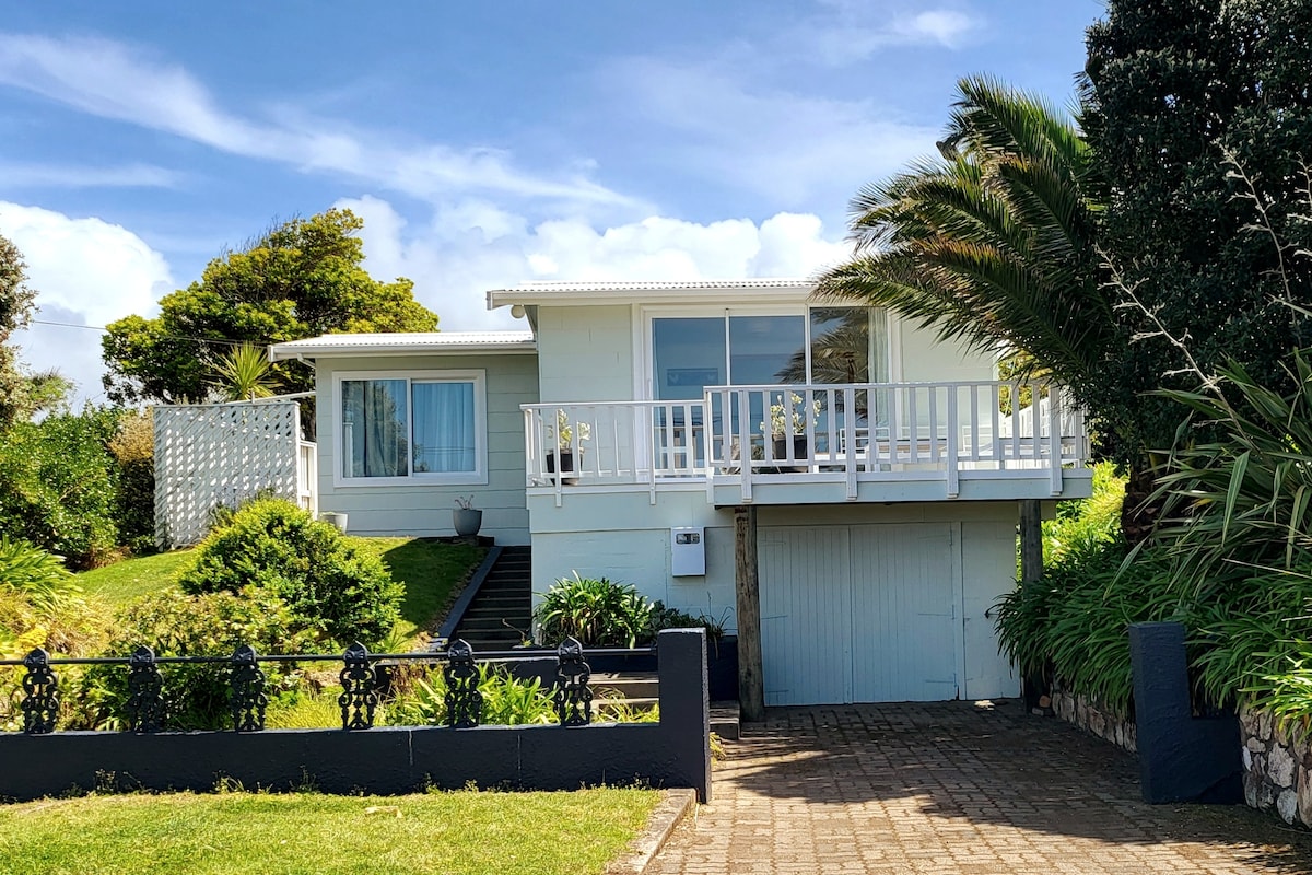 A light-colored two-story house is positioned on a grassy slope, featuring large windows that offer a view of the surroundings. A spacious deck extends from the front of the house, bordered by greenery. The driveway leads to an integrated garage underneath.