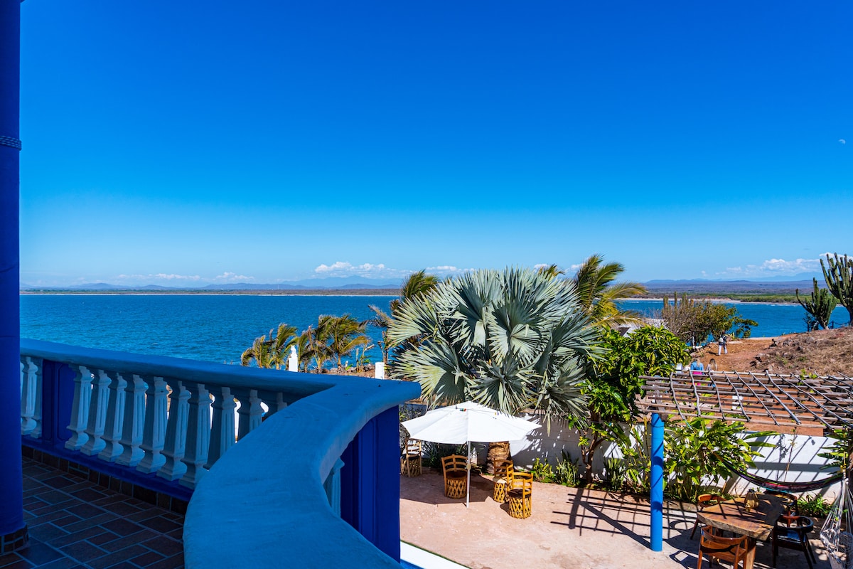 A balcony view highlights the vibrant blue water and distant coastline under a clear sky. Lush palm trees and outdoor furniture are visible, creating a serene outdoor space for relaxation. Bright sunshine enhances the scenic beauty of the surroundings.