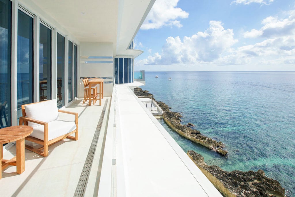 A glass-enclosed balcony offers expansive views of the ocean and sky. Light wooden furniture, including a table and chairs, is positioned along the balcony, while the calm sea stretches out into the distance, accentuated by rocky shoreline.