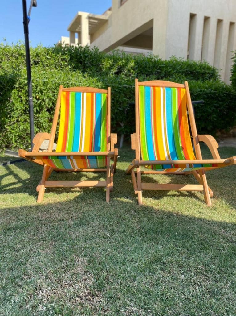 Two wooden lounge chairs with brightly colored striped fabric are positioned on a grassy area, surrounded by neatly trimmed hedges. The outdoor setting is bathed in natural light, creating an inviting atmosphere for relaxation.