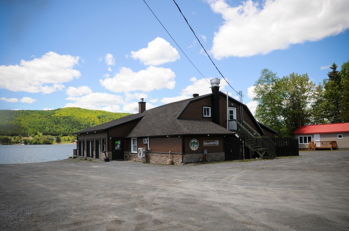 A rustic building is situated along the edge of the water, surrounded by lush greenery and distant hills. The structure features a dark facade with large windows and a sloped roof, complemented by a wooden deck and outdoor stairs. A gravel area is visible in the foreground.