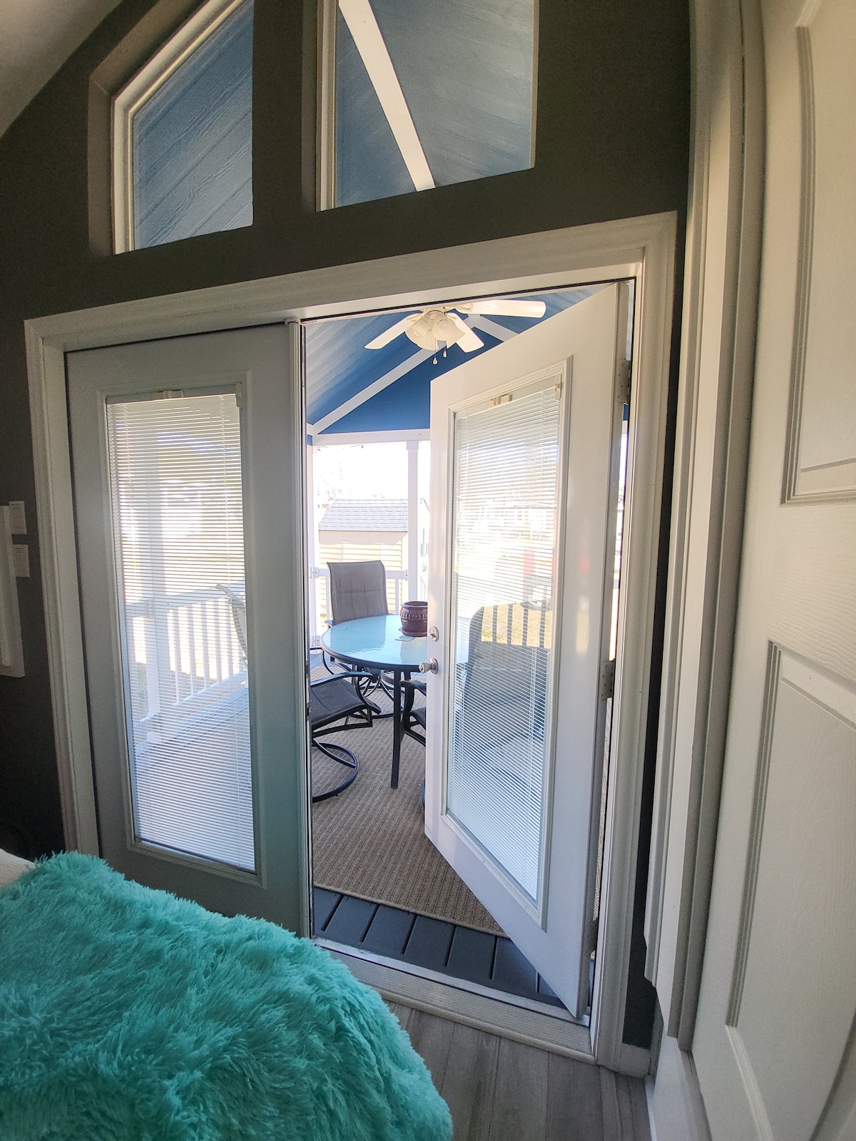 Double doors open to a cozy outdoor seating area, featuring a small round table and four chairs. The space is shaded by a blue ceiling and light streams in through tinted windows, creating an inviting atmosphere.