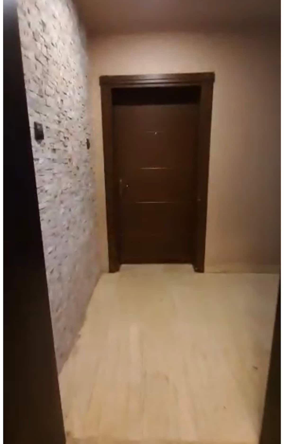 A simple hallway is displayed, featuring smooth stone wall accents alongside a solid brown door. The floor is finished with light-colored tiles, contributing to an open and inviting entrance.