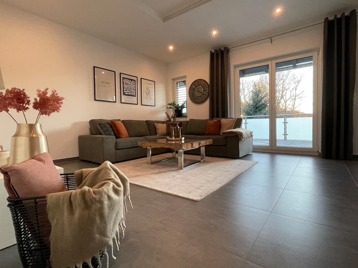 A modern living area features a spacious sectional sofa adorned with colorful cushions. A rustic coffee table is centered on a light area rug. Large glass doors allow natural light to enter, leading to a balcony. Decor accents include artwork and a stylish vase.